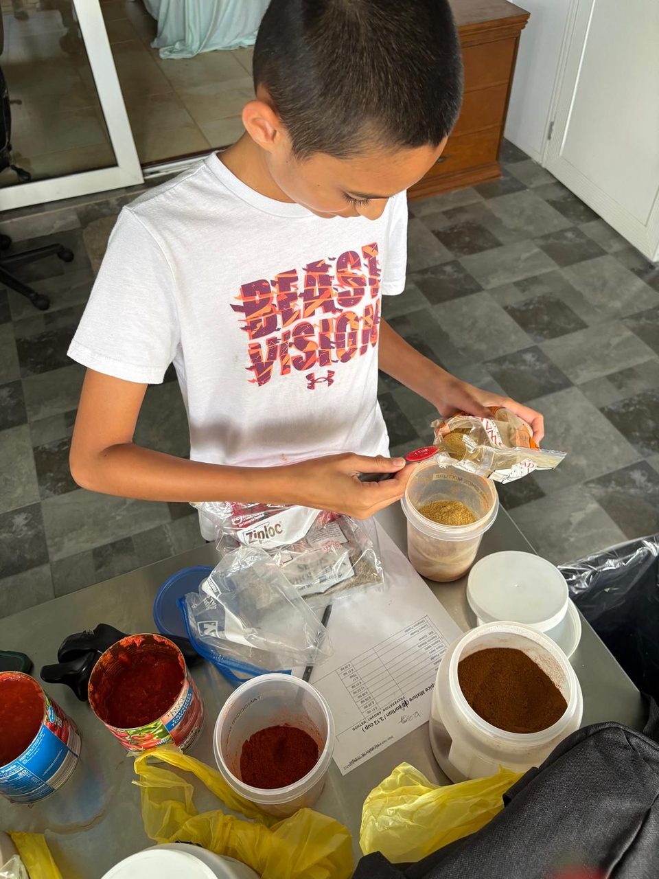 Jian measuring ground spices with a measuring spoon, a printed spice-mixture recipe and containers of red and brown spice powders in front of him