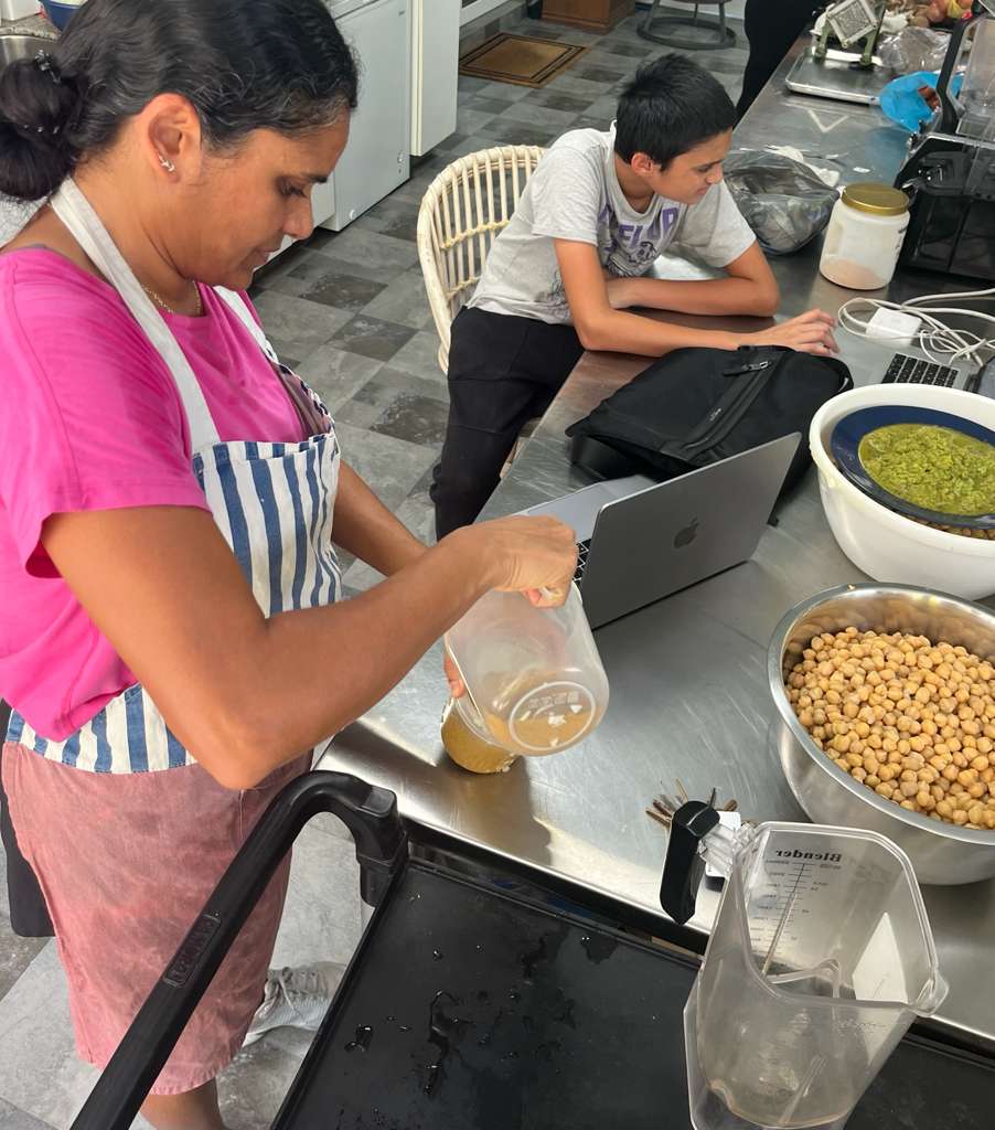 Jilna, in a striped apron, preparing food at the steel kitchen counter beside a bowl of chickpeas, with her son Jian sitting next to her